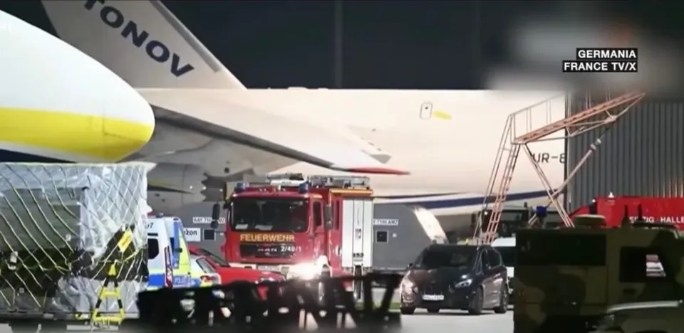 Un avion mare este parcat pe o pistă de noapte lângă un camion de pompieri roșu și un vehicul de serviciu albastru.