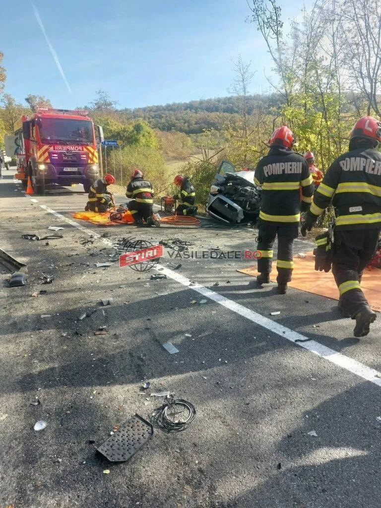 Pompieri în echipament de protecție portocaliu și negru lucrează pe un drum lângă un vehicul de culoare închisă și resturi împrăștiate pe asfalt.