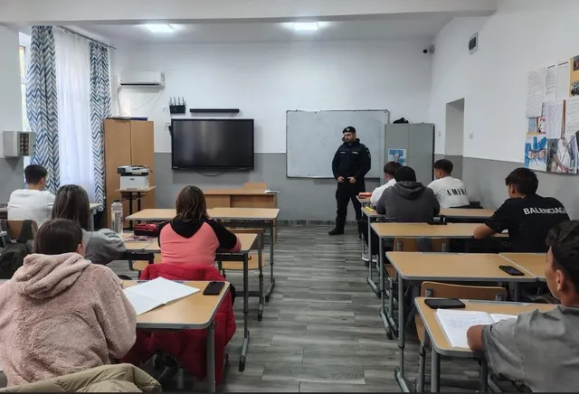 Un bărbat în uniformă neagră stă în fața unui grup de elevi așezați la bănci într-o sală de clasă cu un ecran și un panou alb.