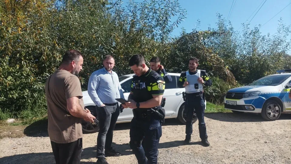 Cinci bărbați stau pe un drum de pietriș lângă două mașini albe și albastre, în fața unor copaci verzi sub un cer senin.
