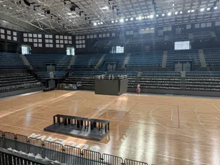 Interiorul unei săli de sport cu parchet deschis, tribune cu scaune negre și o structură neagră situată pe teren.