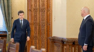 Un bărbat într-un costum închis la culoare stă în picioare lângă o ușă de lemn sculptată, privind spre un alt bărbat cu spatele la cameră.