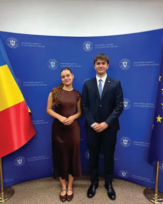 Un bărbat în costum și o femeie în rochie maro stau în picioare în fața unui fundal albastru cu inscripții repetitive.
