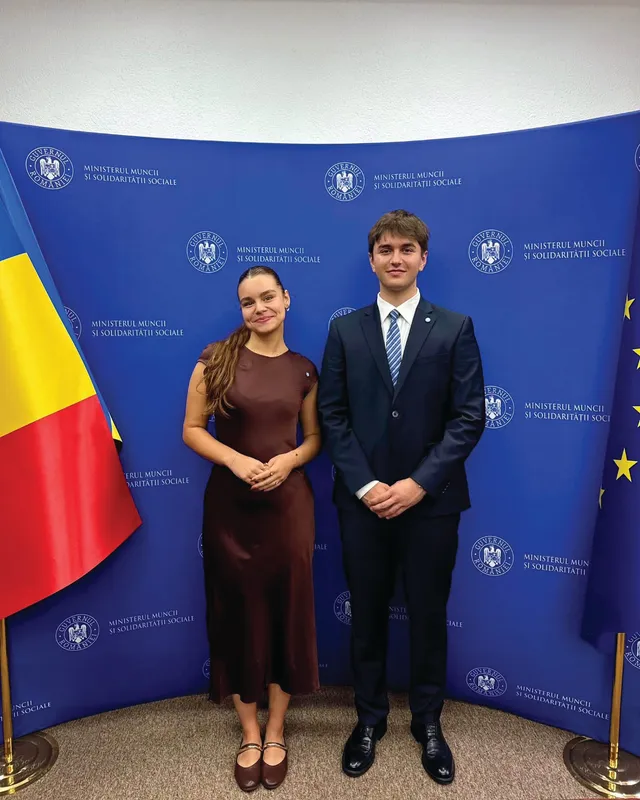 Un bărbat în costum și o femeie în rochie maro stau în picioare în fața unui fundal albastru cu inscripții repetitive.