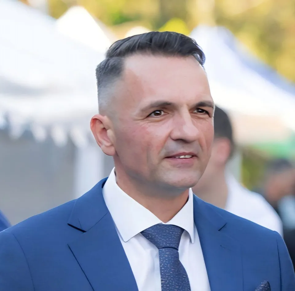 Un bărbat cu părul scurt și coafat pe o parte poartă un sacou albastru, cămașă albă și cravată închisă la culoare.