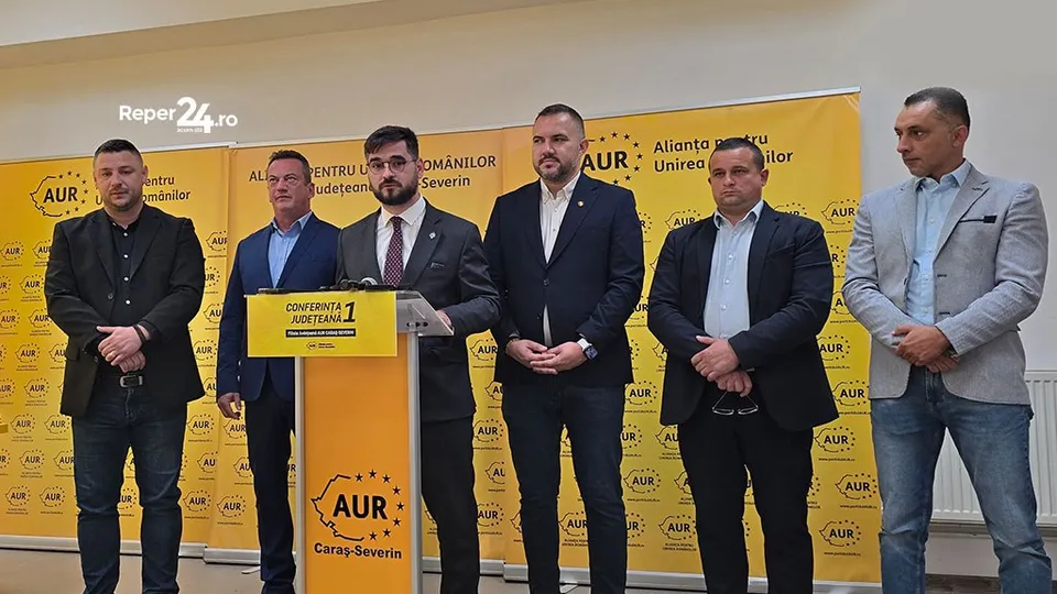 Șase bărbați stau în picioare în fața unui panou galben cu text și logo-uri, unul dintre ei fiind poziționat în spatele unui podium.