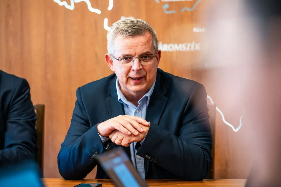 Un bărbat cu ochelari și sacou închis peste o cămașă deschisă stă la o masă cu mâinile împreunate.