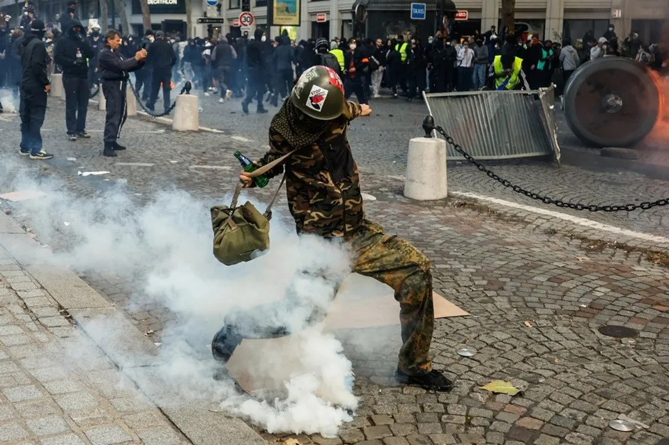 O persoană îmbrăcată în haine de camuflaj și cască aruncă un obiect într-un nor de fum pe un drum pavat.
