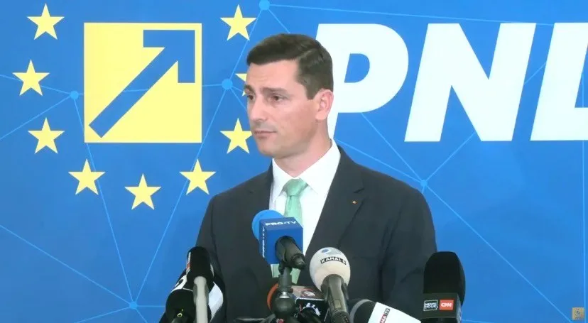 Un bărbat cu sacou închis și cravată deschisă stă în fața unui fundal albastru cu un logo galben și text alb lângă microfoane.
