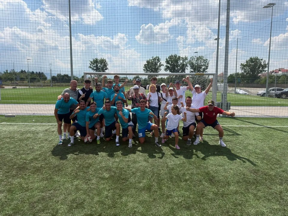 Un grup de oameni îmbrăcați în echipamente sportive stau grupați pe un teren de fotbal cu iarbă artificială sub un cer înnorat.