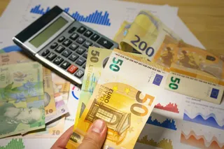 O mână ține bancnote de euro pe un fundal format din un calculator, diverse bancnote și grafice pe hârtie.