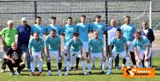 O echipă de fotbal masculin, îmbrăcată în tricouri albastre deschis și pantaloni albi, pozând grupat pe un teren de gazon cu un poartă de fundal.