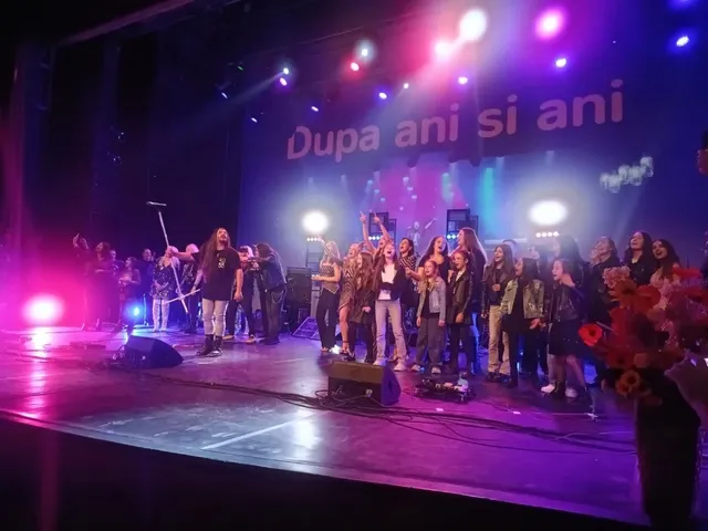 Un grup de persoane stă pe o scenă iluminată de lumini colorate sub un ecran mare cu textul scris în fundal.