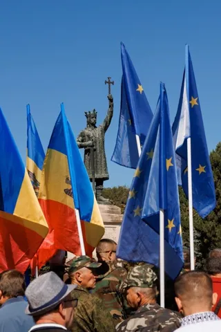 O mulțime de oameni, unii în uniforme militare, stau în fața unui monument, fluturând steaguri ale Republicii Moldova și ale Uniunii Europene.
