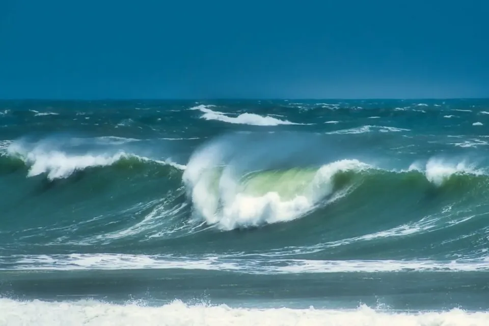 Valuri mari de culoare verde închis cu spumă albă se ridică pe un ocean sub un cer albastru.