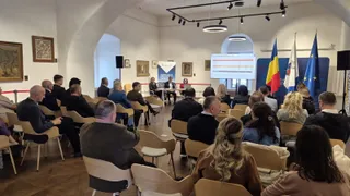 O grupă de persoane stă pe scaune într-o încăpere cu pereți albi, orientată spre un grup de oameni așezați la o masă sub un ecran.