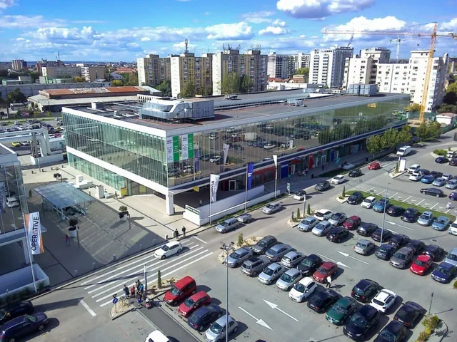Imagine aeriană cu o clădire modernă de sticlă, parcări pline, străzi, câteva persoane și blocuri în fundal.