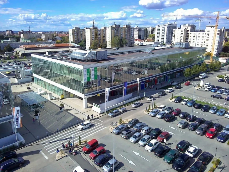 Imagine aeriană cu o clădire modernă de sticlă, parcări pline, străzi, câteva persoane și blocuri în fundal.
