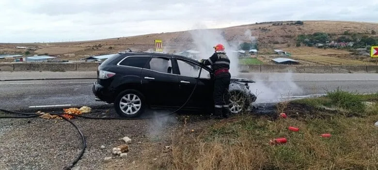 Un pompier cu cască portocalie stă lângă un autoturism negru care scoate fum pe marginea unui drum asfaltat.