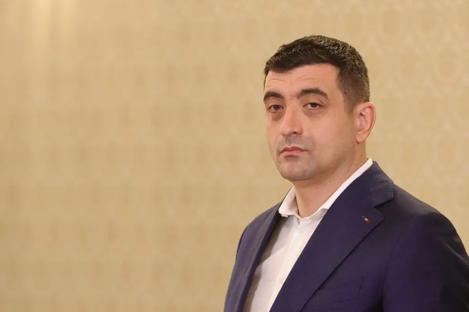 Un bărbat cu părul scurt și întunecat poartă un sacou albastru peste o cămașă albă, stând în fața unui perete cu model discret.