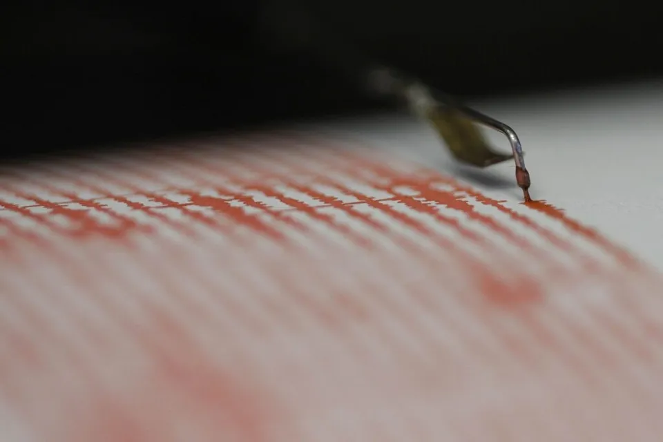 Un pen de seismograf trasează linii ondulate de culoare roșie pe o suprafață albă sub o lumină difuză.