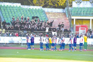 Un grup de fotbaliști în echipamente sportive colorate stau pe teren, în timp ce mulți spectatori îmbrăcați în negru stau pe tribunele unui stadion.