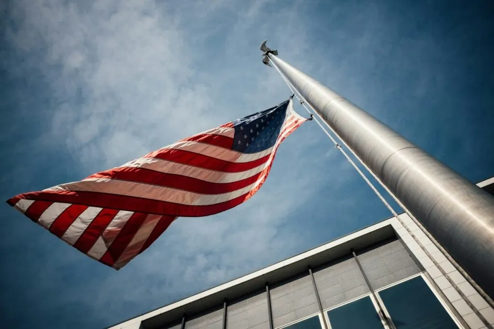 Un steag american flutură pe un catarg înalt, cu o clădire modernă și un cer albastru cu nori în fundal.