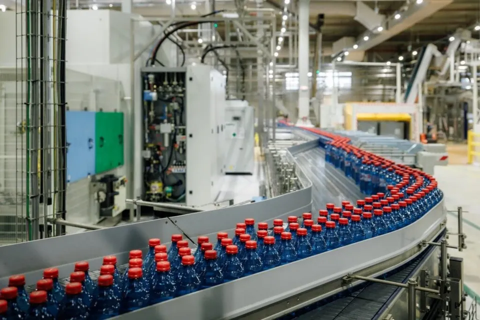 Linii de producție cu sticle de apă albastre cu capace roșii care se deplasează pe o bandă de transport într-o fabrică cu utilaje industriale mari.