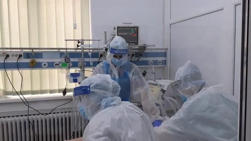 Persoane purtând echipament de protecție alb și măști fac parte dintr-un cadru medical cu echipamente de monitorizare și dispozitive medicale în fundal.