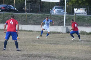 Trei jucători de fotbal sunt pe un teren de iarbă uscată, unul cu tricou albastru cu dungi verticale și celălalt doi cu tricouri roșii.