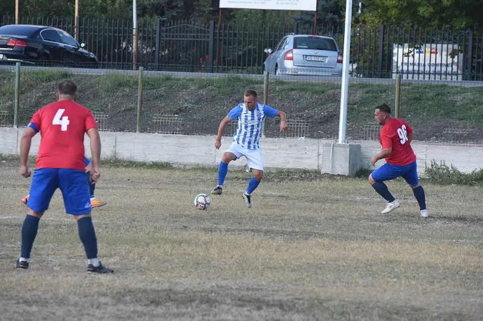 Trei jucători de fotbal sunt pe un teren de iarbă uscată, unul cu tricou albastru cu dungi verticale și celălalt doi cu tricouri roșii.