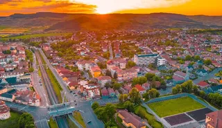 Aerial view of the town of Blaj at sunset, showing houses, streets, green areas, and hills in the background under an orange sky.