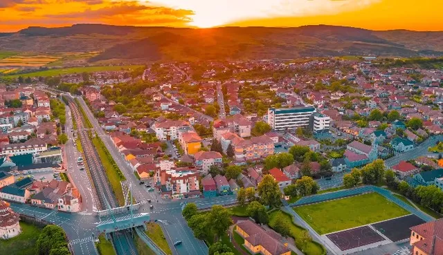 Aerial view of the town of Blaj at sunset, showing houses, streets, green areas, and hills in the background under an orange sky.