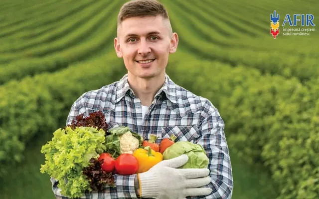 Un bărbat zâmbind poartă o cămașă în carouri și ține în brațe diverse legume, inclusiv salată, conopidă și ardei, într-un câmp verde.