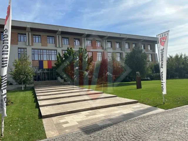 O clădire modernă cu un pavaj din piatră pe un i verde, flancată de steaguri, cu o arhitectură simetrică.