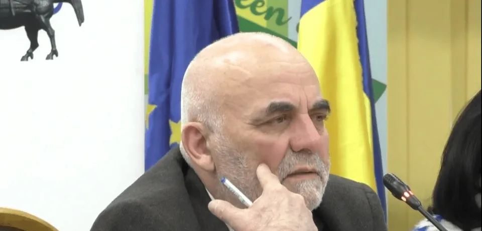 Un bărbat cu părul scurt și barbă căruntă stă la o masă cu mâna la față, având steaguri în fundal.