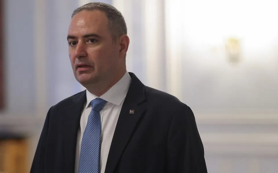 Un bărbat cu părul scurt, purtând sacou închis la culoare, cămașă albă și cravată albastră cu puncte mici, privește spre lateral într-un decor interior.