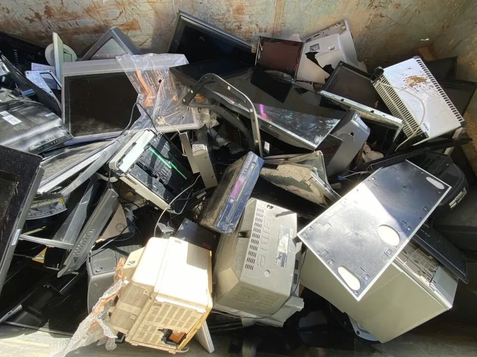 Un amestec de monitoare, carcase de calculatoare și diverse componente electronice de diverse culori este depozitat într-un recipient metalic mare.