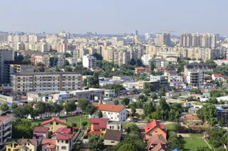 O vedere panoramică asupra unui oraș cu numeroase clădiri de apartamente, case cu acoperiș înclinat și zone verzi cu copaci sub un cer senin.