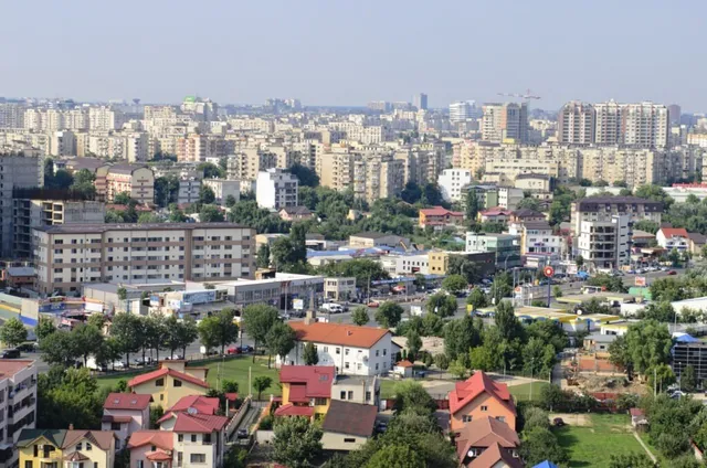 O vedere panoramică asupra unui oraș cu numeroase clădiri de apartamente, case cu acoperiș înclinat și zone verzi cu copaci sub un cer senin.