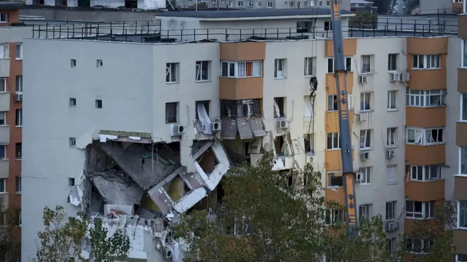 O cameră de tip macarica este poziționată lângă un bloc de apartamente care prezintă o porțiune de fațadă și etaje distruse.