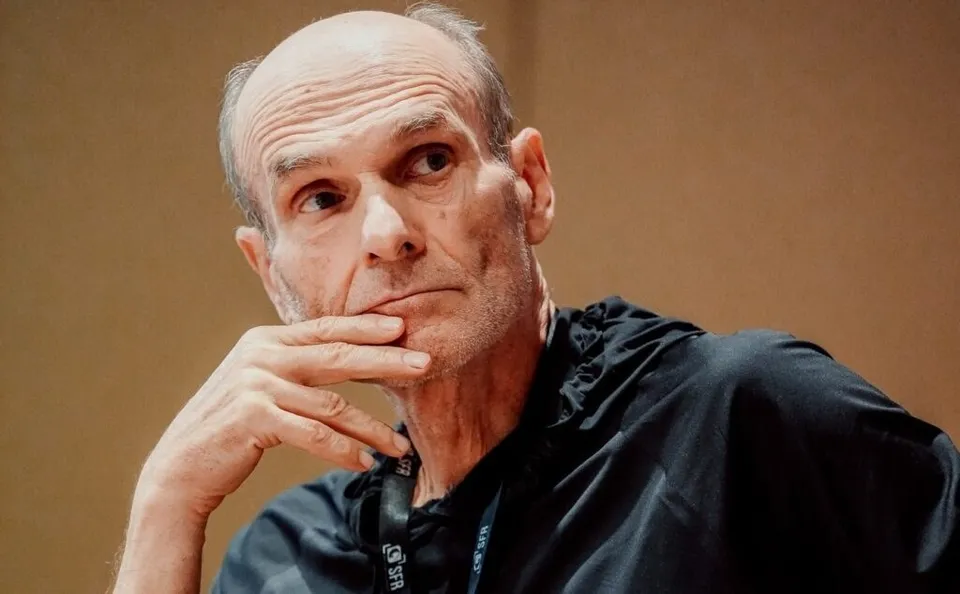 Un bărbat cu părul rare și cămașă neagră stă cu mâna la bărbie, privind spre lateral.