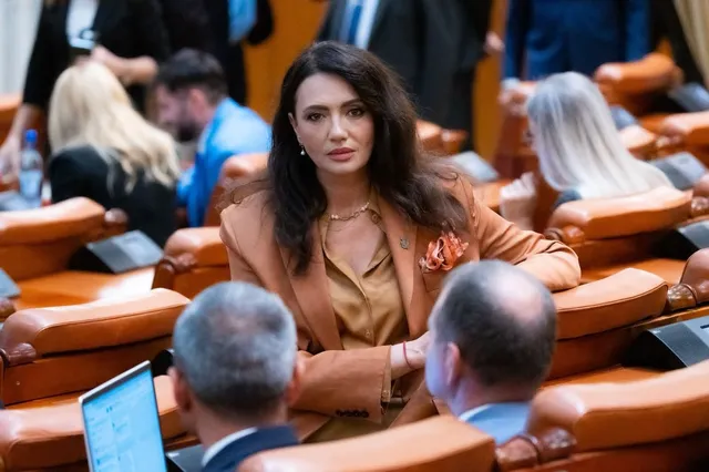O femeie cu părul închis la culoare, purtând o jachetă maro și un colier, stă într-o sală cu scaune de lemn și oameni în fundal.