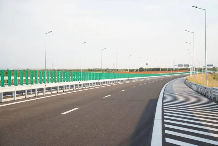 O autostradă cu asfalt negru, linii albe marcate pe carosabil și garduri metalice de protecție, sub un cer deschis și polei de iluminat stradal.