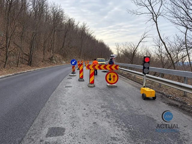 O bandă de rulare este blocată de conuri de semnalizare portocalii și un semn de circulație cu săgeți pe un drum asfaltat lângă un gard metalic.