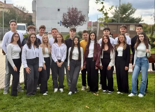 Un grup de tineri îmbrățișați într-un șir, purtând bluze albe și pantaloni gri sau negri, pozând pe un peluz verde.