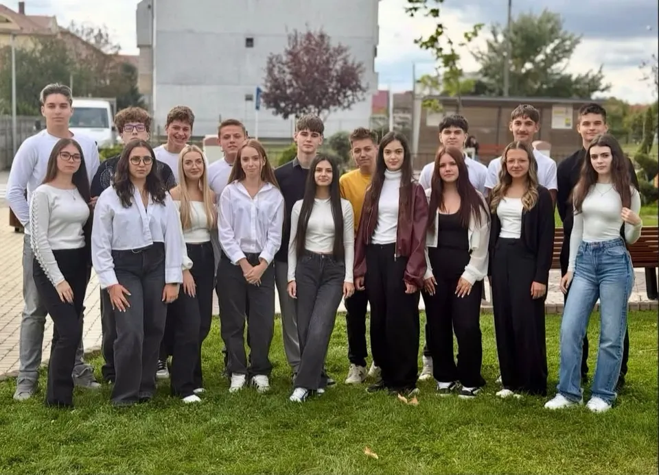 Un grup de tineri îmbrățișați într-un șir, purtând bluze albe și pantaloni gri sau negri, pozând pe un peluz verde.