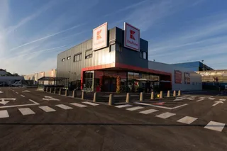 O clădire comercială gri cu semne Kaufland este situată lângă un parcare asfaltat cu marcaje albe pe sol sub un cer senin.