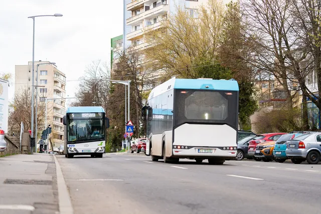 Un autobuz alb și albastru circulă pe un drum asfaltat lângă o linie de mașini parcate și clădiri înalte sub un cer înnorat.