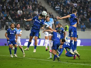 Câțiva jucători de fotbal în echipamente albastre și albe concurează pentru un balon în aer pe un teren de fotbal verde.